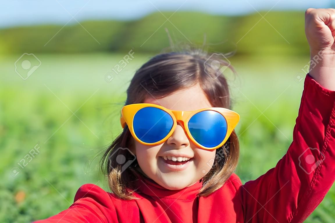 Glückliches kleines Mädchen mit großen Sonnenbrillen auf der Wiese zu Fuß und in die Sonne schaut. Mädchen mit den Händen in der Luft machen Daumen nach oben