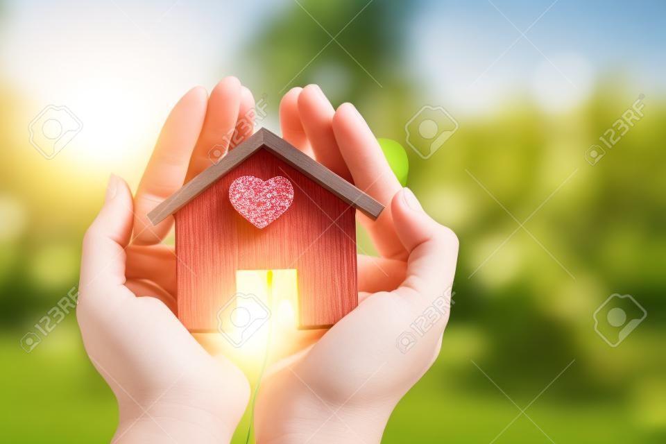 Mão de mulher segurando um modelo de casa, coloque um coração vermelho à luz do sol no parque público, empréstimos para imóveis ou economize dinheiro para comprar uma nova casa para a família no conceito futuro.