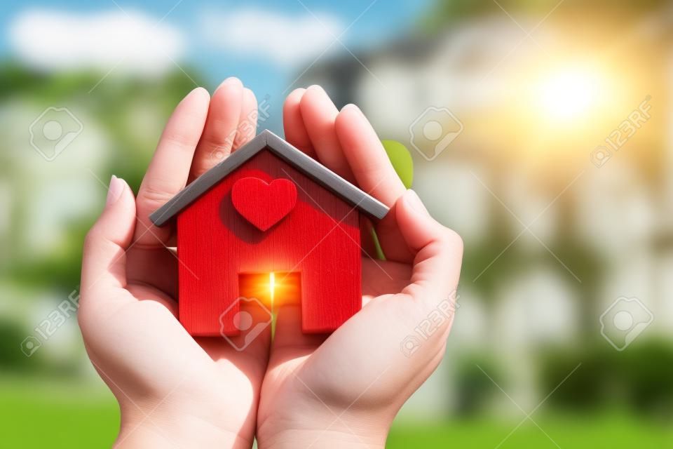 Woman hand holding a home model stick a red heart in the sunlight in the public park, Loans for real estate or save money to buy a new house for family in the future concept.