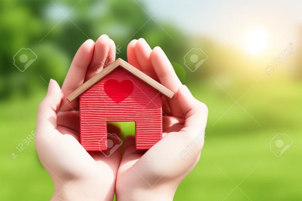 拿著一個家庭模型的婦女手在陽光下的一個紅色心臟在公園，貸款為房地產或在未來概念的存金錢為家庭買一個新的房子。