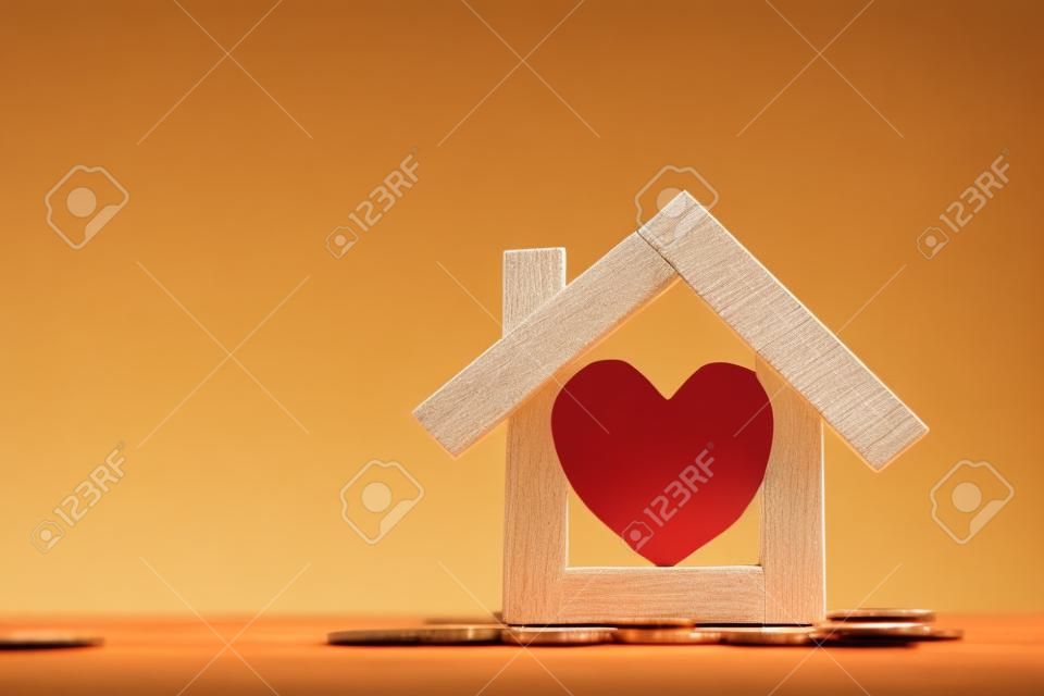 Rotes Herz in der Mitte des Holzhausmodells legte auf die Goldmünze mit wachsendem Interesse an Sonnenlicht im öffentlichen Park, Unternehmensinvestitionen oder Geld sparen, um Immobilien für Familienkonzepte zu kaufen.