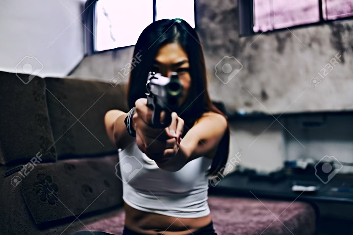 Donna asiatica con in mano una pistola, che punta alla telecamera Nell'atmosfera della vecchia stanza, fatiscente, pericolosa, fuorilegge, violenta, barbara