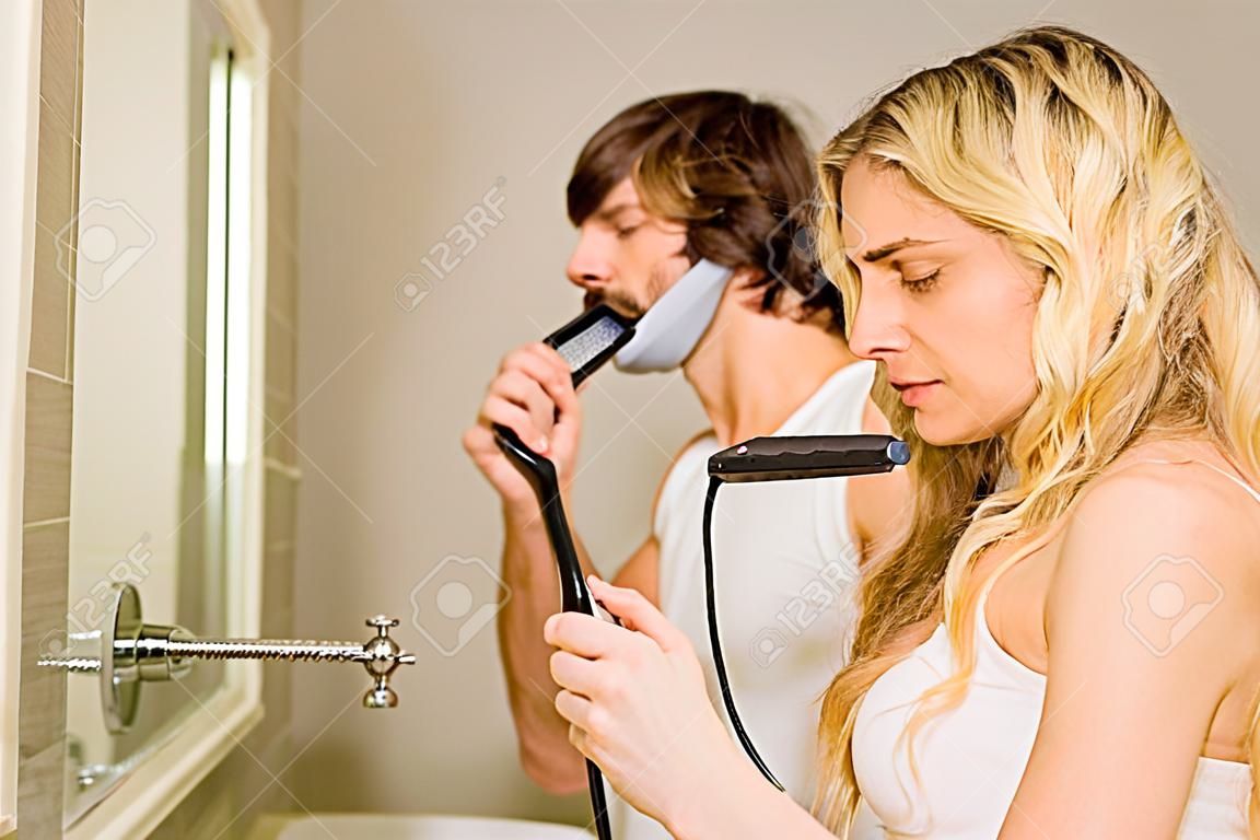Cute couple using a razor and a straightener in the bathroom at home