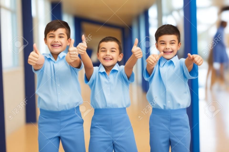 Portret uśmiechniętych dzieci w wieku szkolnym pokazujących kciuki do góry na korytarzu w szkole