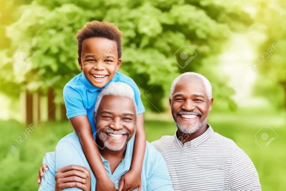 Porträt eines lächelnden afroamerikanischen Großvaters im Garten mit seinem erwachsenen Sohn und seinem Enkel auf dem Huckepack.