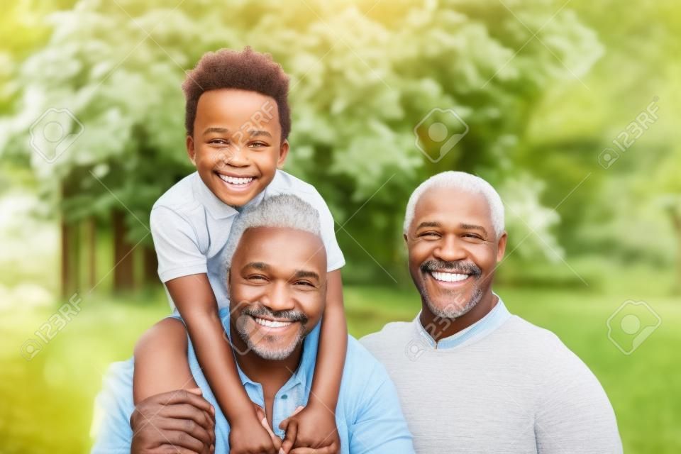 Porträt eines lächelnden afroamerikanischen Großvaters im Garten mit seinem erwachsenen Sohn und seinem Enkel auf dem Huckepack.