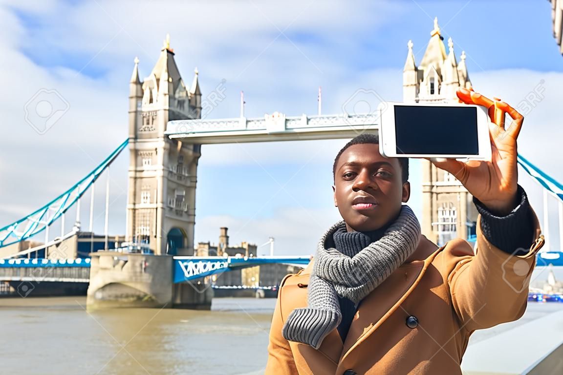 Smiling black man taking selfie in London with Tower Bridge on background. 