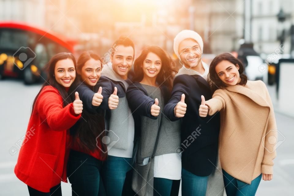 Successful group of friends showing thumbs upin London. They are four women and two men in their twenties, they are standing in a row, all very close by each other. Friendship and lifestyle concepts.