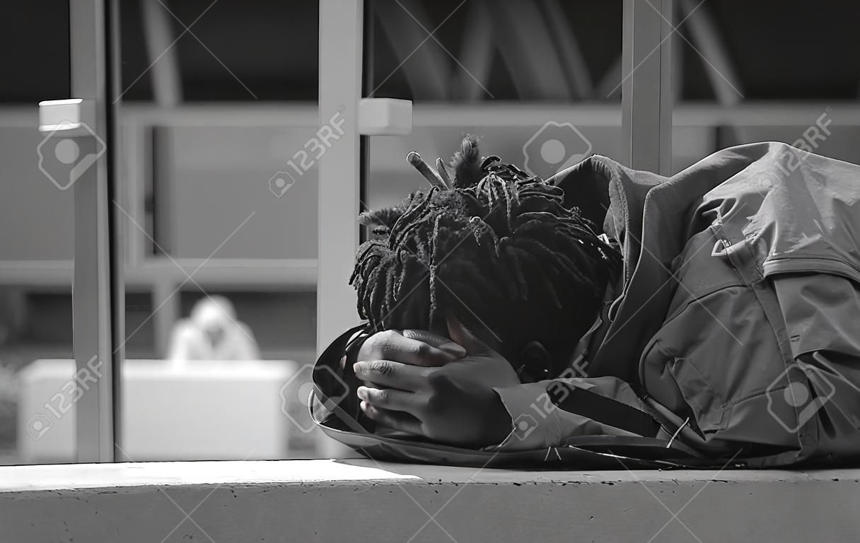 Ujęcie w skali szarości przedstawiające Afroamerykanina w płaszczu i leżącego na ławce na zewnątrz