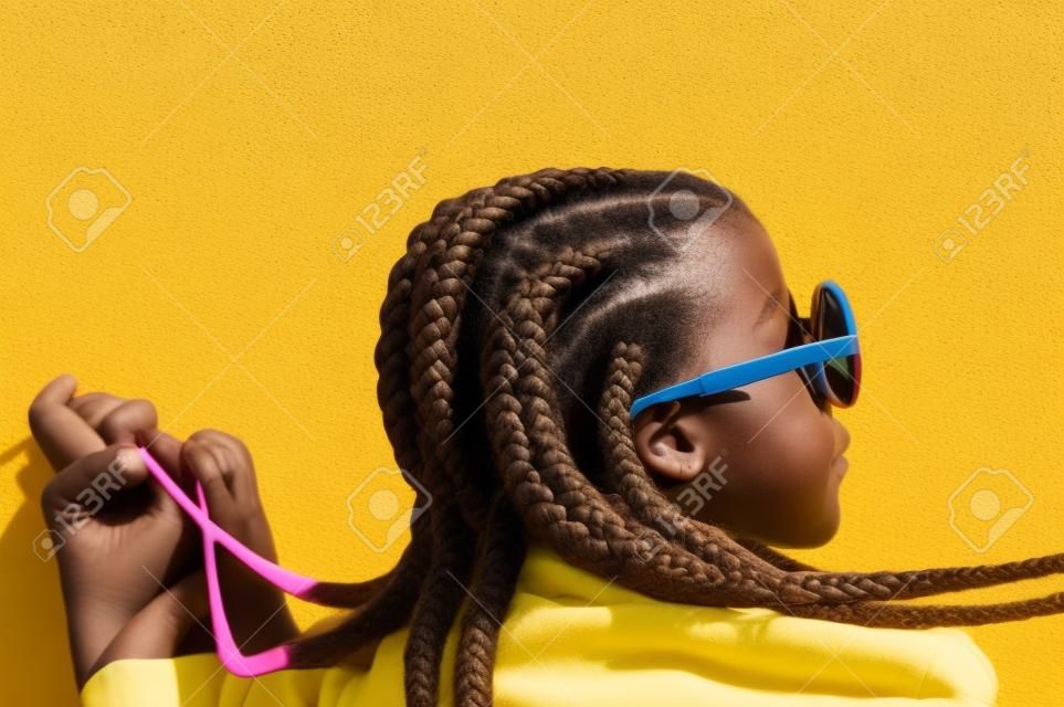 Una giovane ragazza con trecce africane, occhiali da sole e cappotto rosa davanti a un muro giallo