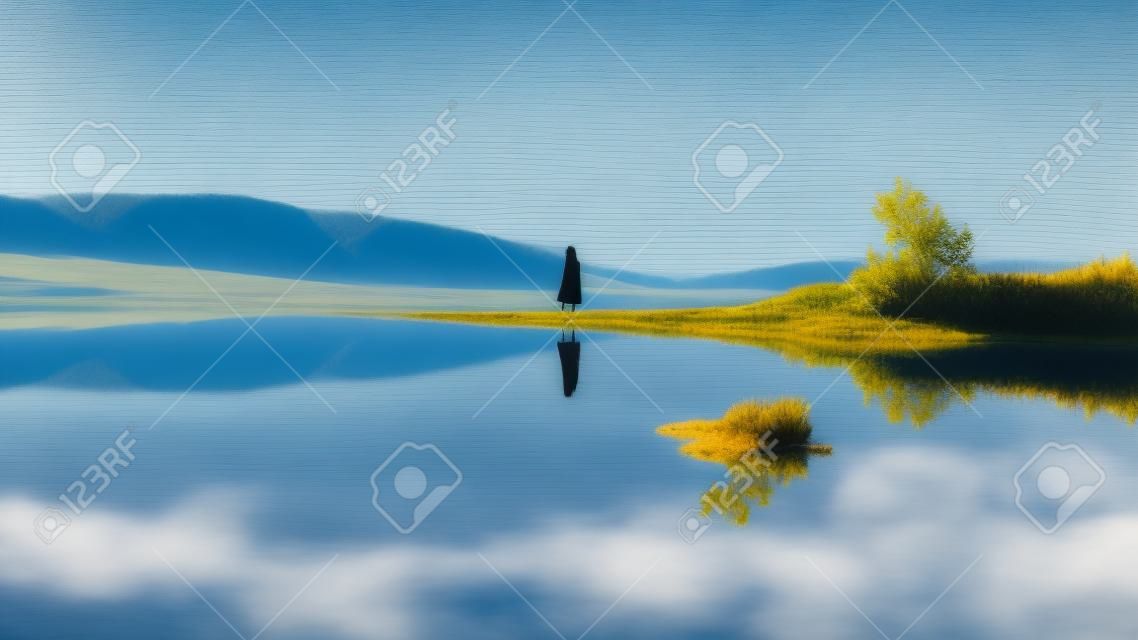 A scenic view of a woman's reflection in the calm lake under a clear sky background