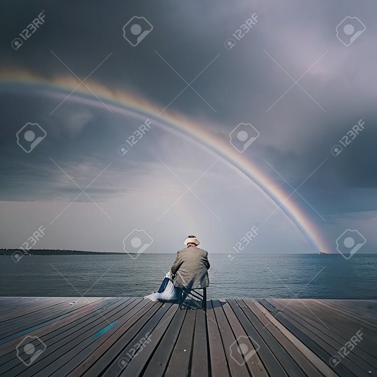 An old male sitting on a pier in the sea with a rainbow in the background sky