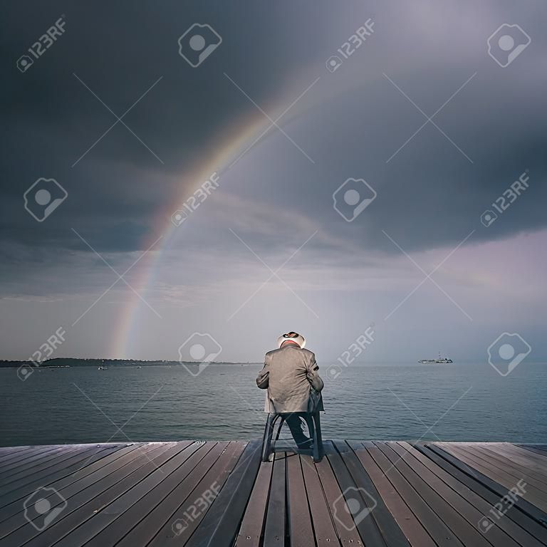 An old male sitting on a pier in the sea with a rainbow in the background sky