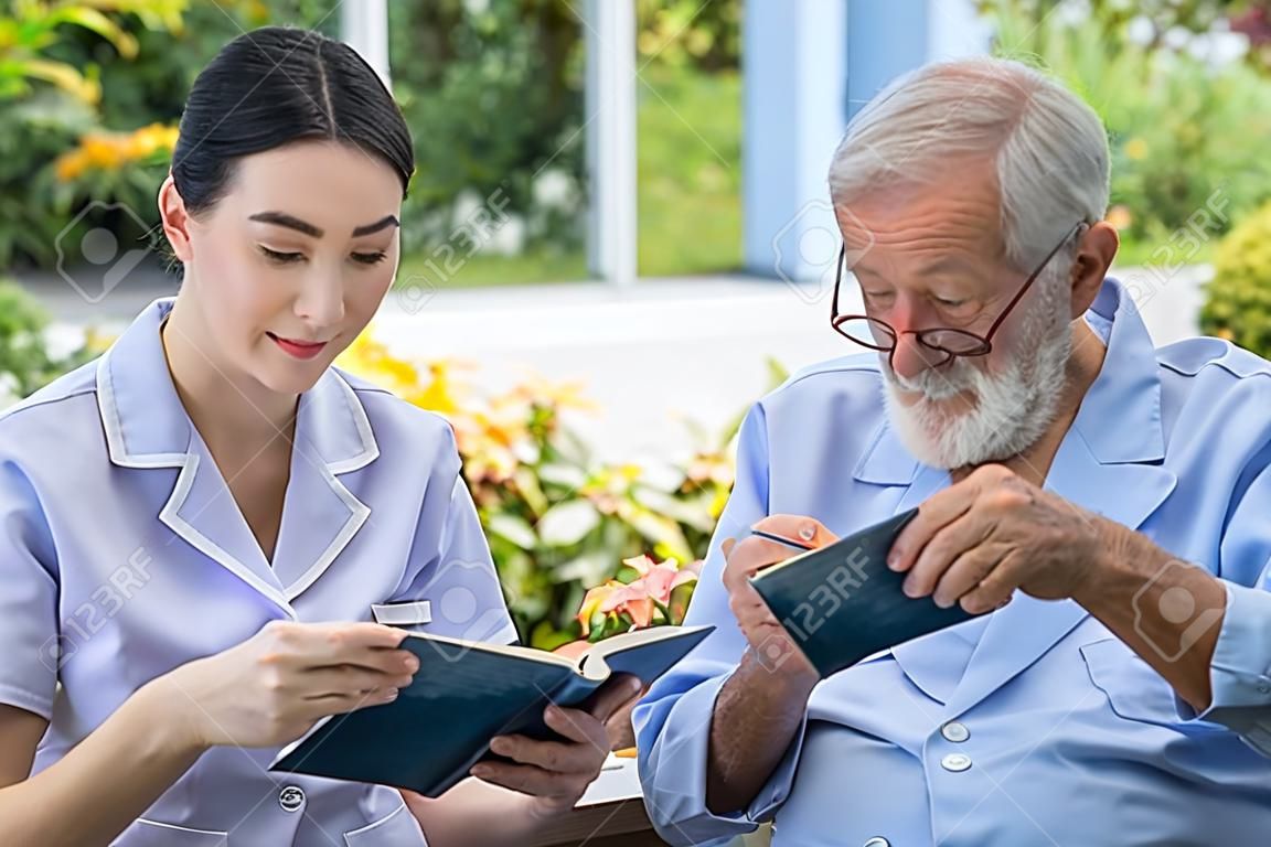 Älterer älterer Mann liest Buch mit Krankenschwester beim Frühstück im Garten im Pflegeheim with