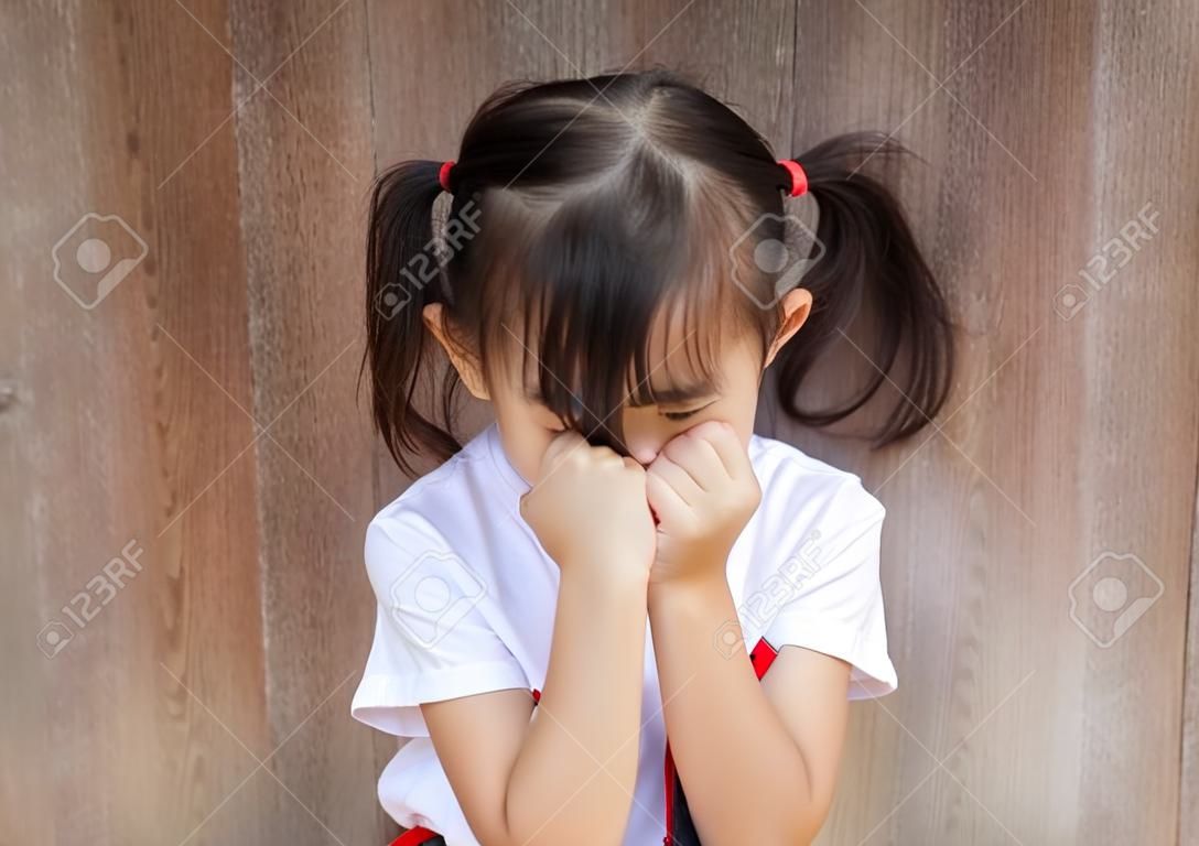 Retrato de medio cuerpo de una adorable niña asiática, de unos 4 años con pantalones blanco, que está posando con cara descarada, llorando, jugando con la cámara en el fondo borroso de una puerta de madera antigua.