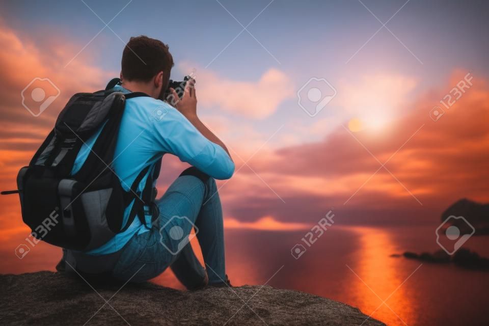 Young european male tourist with a backpack sitting at the cliff by the sea and taking photo of beautiful sunset by his retro camera. Mock up, copy space for your text.