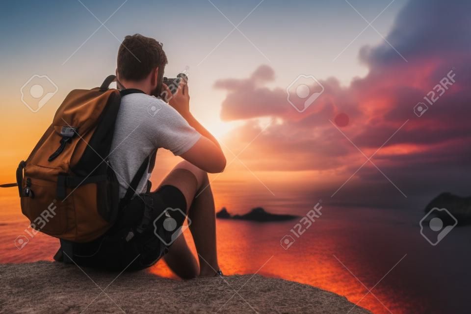 Young european male tourist with a backpack sitting at the cliff by the sea and taking photo of beautiful sunset by his retro camera. Mock up, copy space for your text.