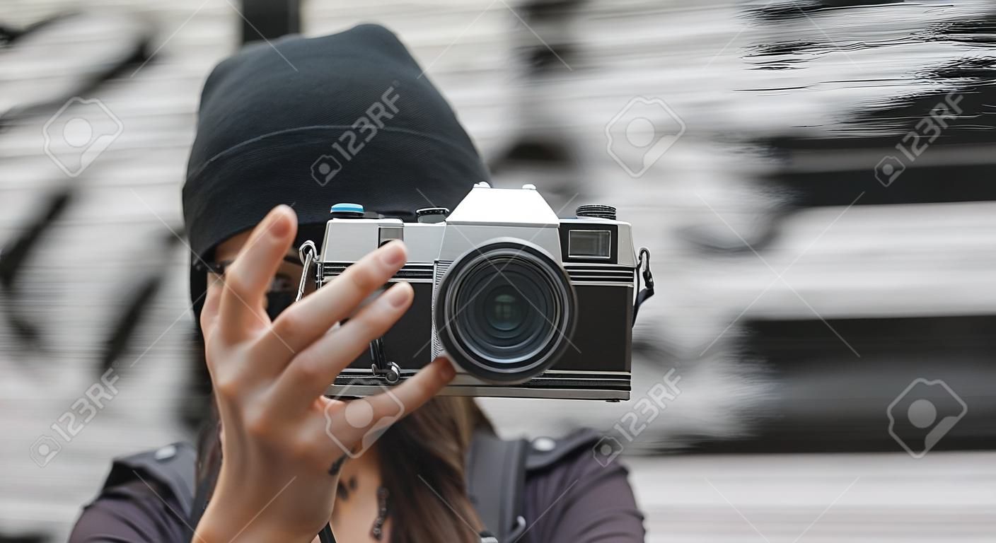 Geernteter Schuss des weiblichen gekleideten gekleideten Touristen mit einem Rucksack , der mit Retro- Filmkamera vor der Graffitiwand aufwirft . Reise- und freiberuflich Konzept . Kopieren Sie Platz für Ihren Text