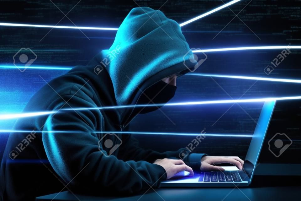 Hacker à capuchon masculin avec un visage caché accédant à des informations personnelles sur un ordinateur portable dans l'obscurité. Concept technologique, cybercriminalité.