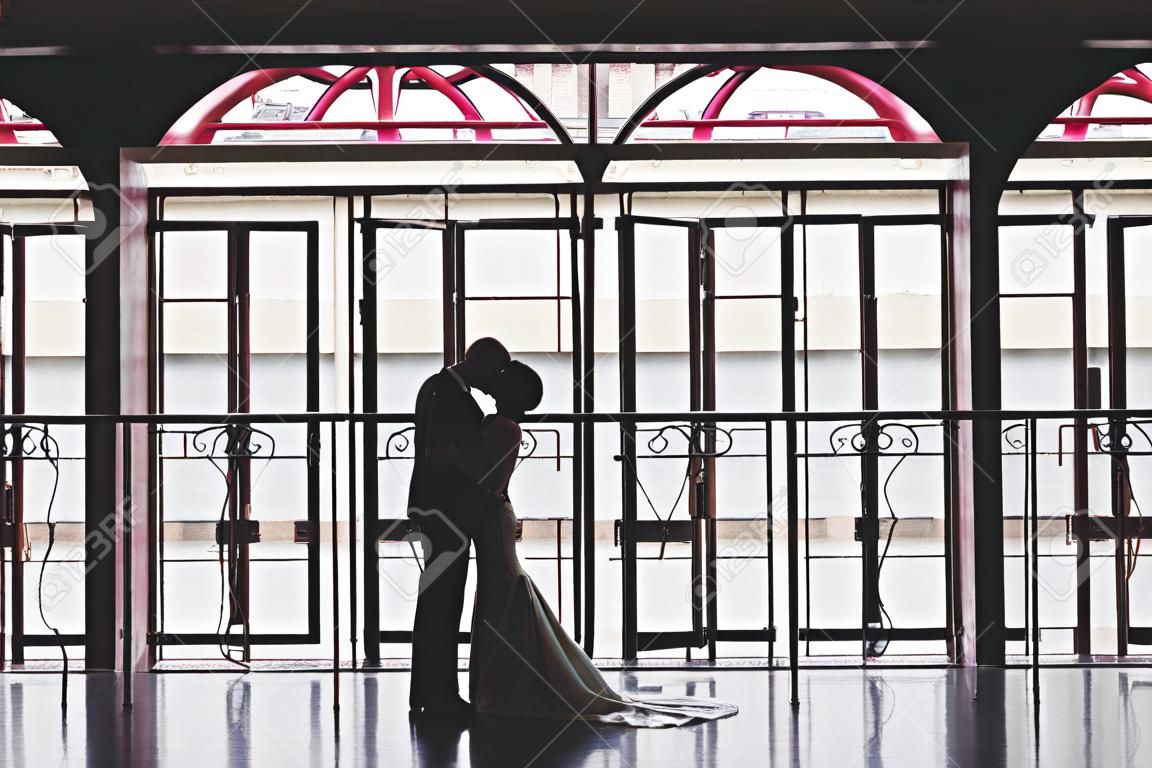 Silhouettes d'un homme en costume et d'une femme en robe et avec un bouquet de fleurs s'embrassant et s'embrassant près d'un grand vitrail panoramique dans un bel intérieur vide