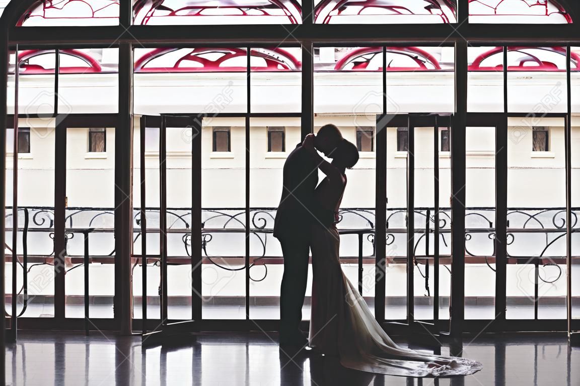 Silhouettes of a man in a suit and a woman in a dress and with a bouquet of flowers kissing and hugging each other near a large panoramic stained glass window in a beautiful empty interior