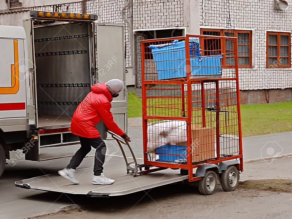 Two workers are loading a trolley with a cage for transporting goods onto the lift of a truck for transportation. Automation of manual work of loaders. Elevator platform on the rear side of the road trailer body. Raise up.