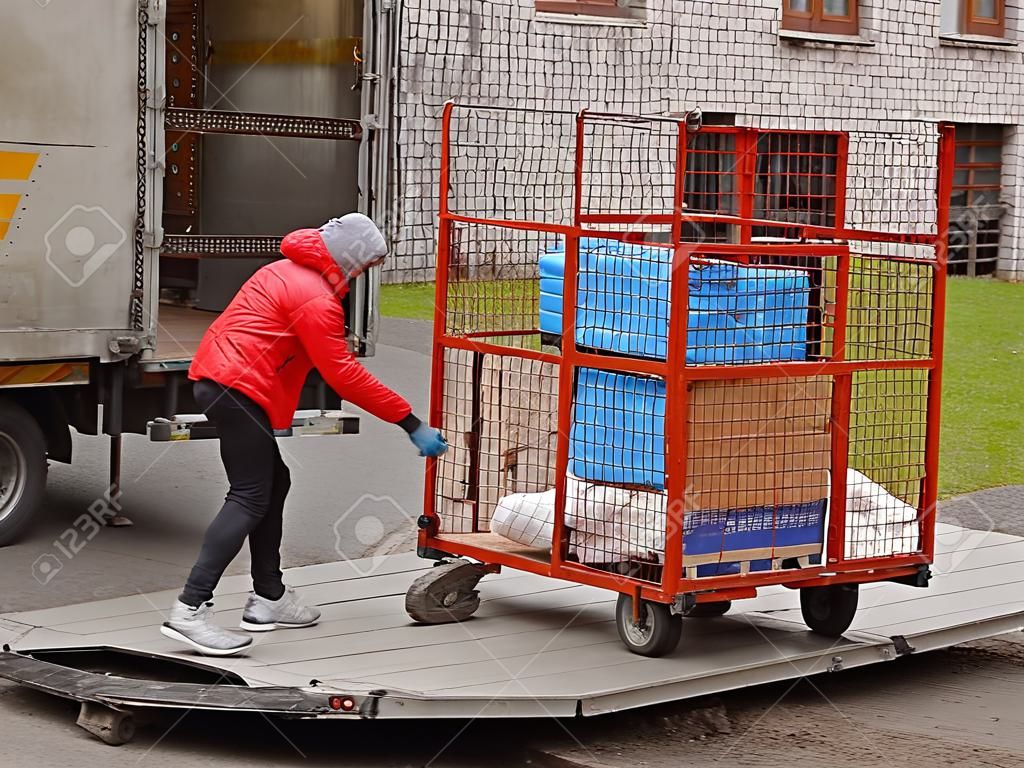 Two workers are loading a trolley with a cage for transporting goods onto the lift of a truck for transportation. Automation of manual work of loaders. Elevator platform on the rear side of the road trailer body. Raise up.
