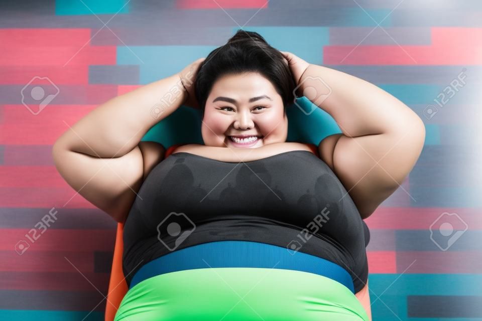 Hohe Betrachtungswinkel einer asiatischen fetten Frau, die in die Kamera lächelt, während sie Bauch-Sit-ups im Fitnesscenter macht