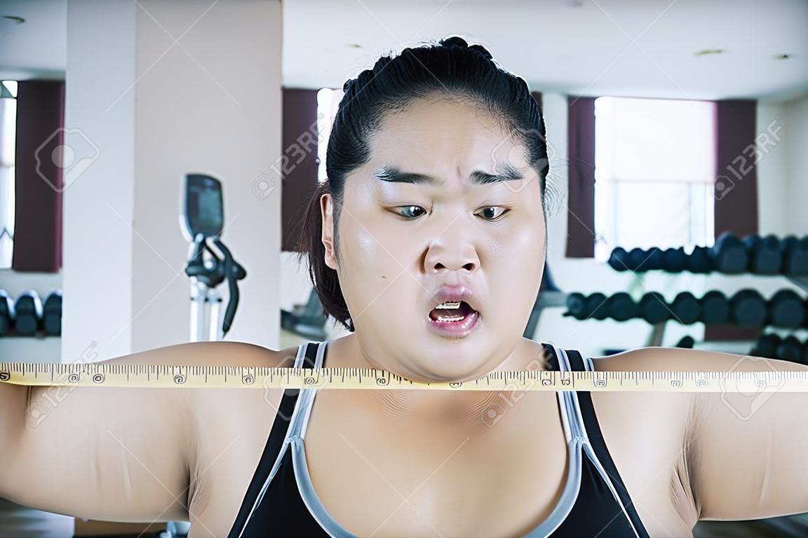 Mujer asiática gorda usando ropa deportiva mientras se siente sorprendida después de ver su chequeo corporal en el gimnasio