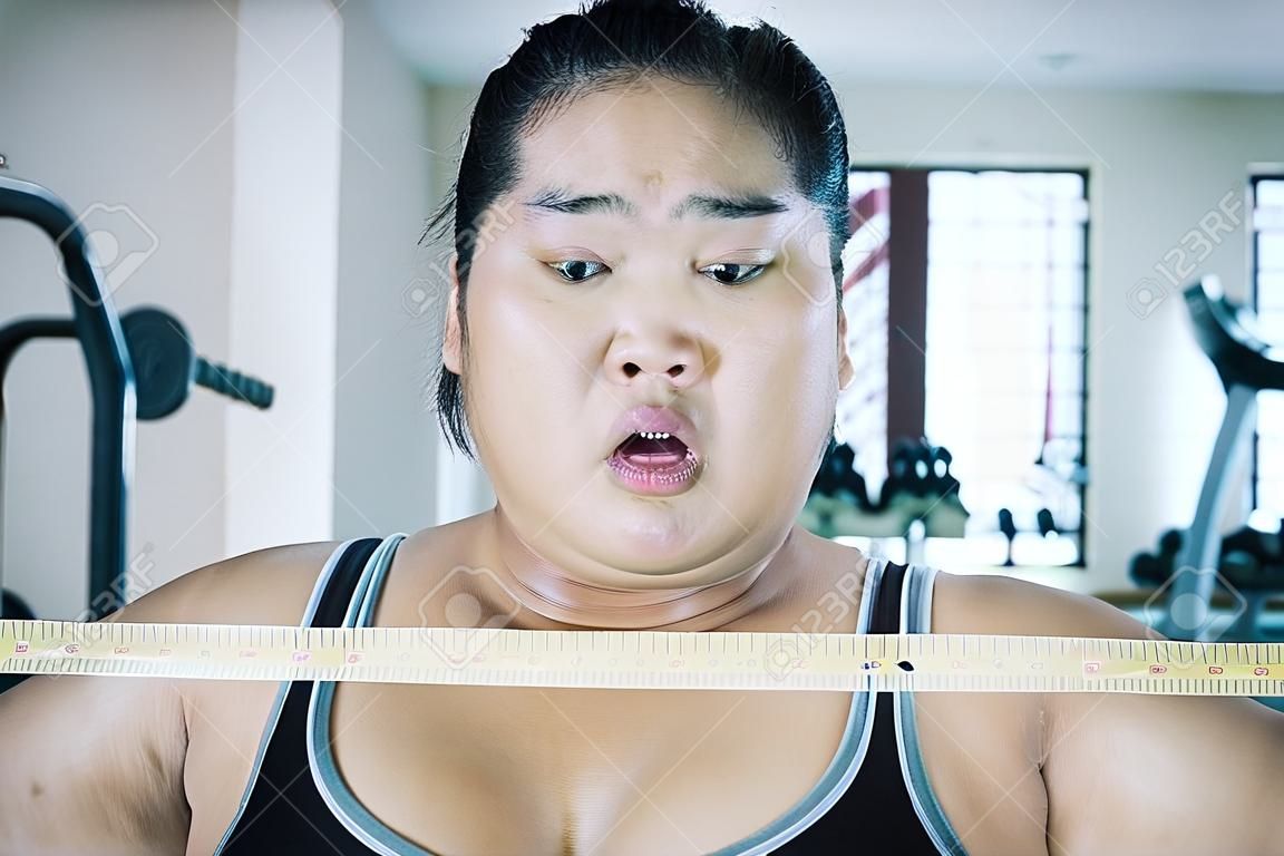 Mujer asiática gorda usando ropa deportiva mientras se siente sorprendida después de ver su chequeo corporal en el gimnasio