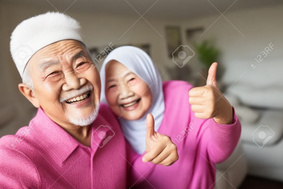 거실에서 셀카를 찍는 동안 엄지손가락을 치켜드는 아시아계 이슬람 노인 부부