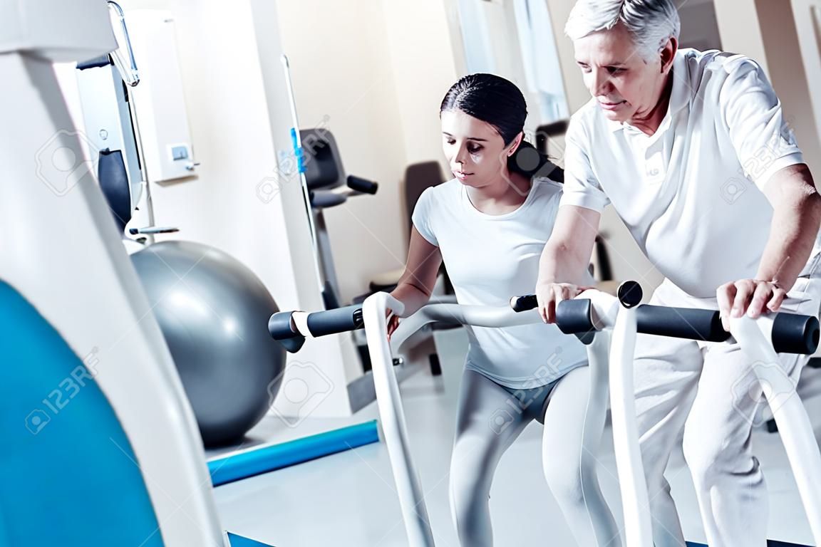 Determined old wrinkled grey-haired man exercising on a training device while a young dark-haired woman smiling and looking at him and touching his leg