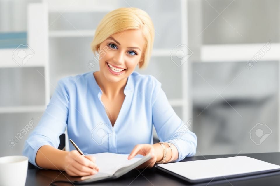 Trabajador ejecutivo. Rubia confiada con una sonrisa en su rostro y abriendo su cuaderno mientras mira directamente a la cámara