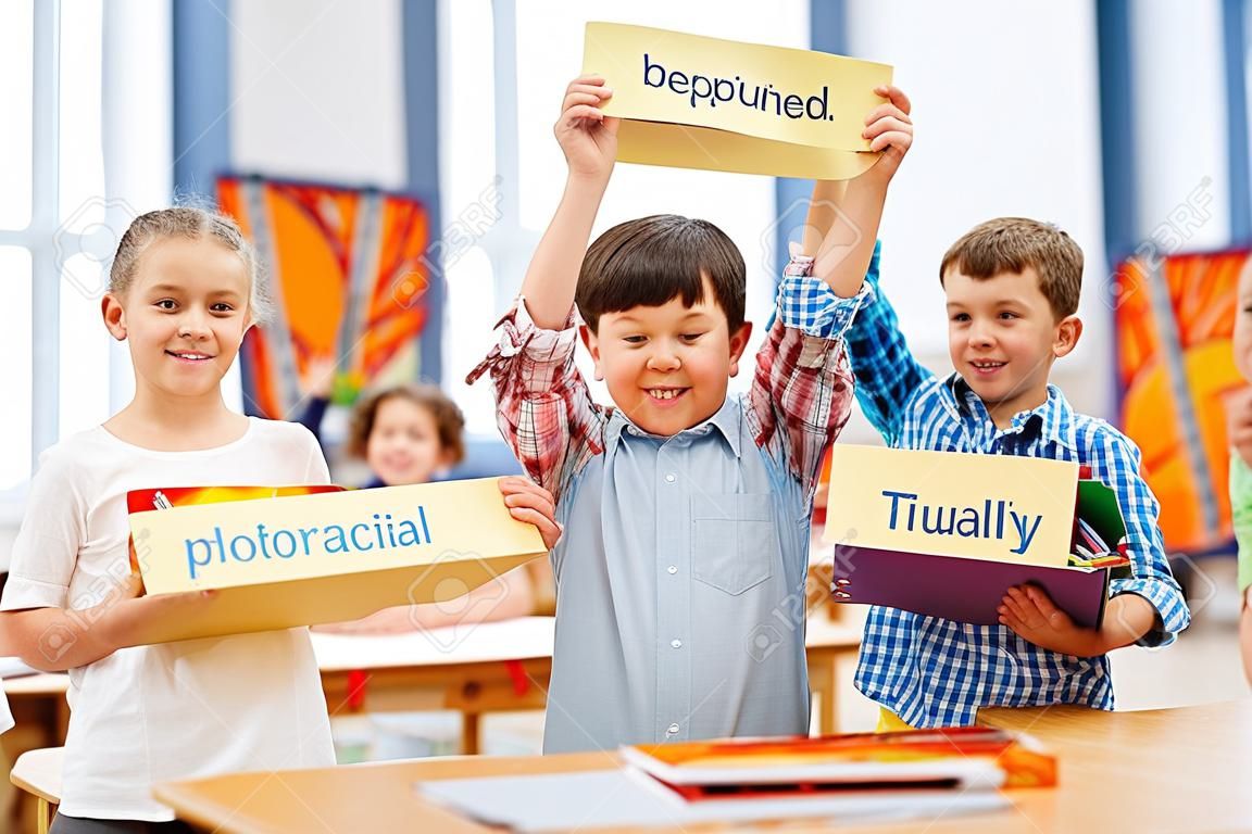 Studying time. Smiling joyful schooldchildren standing in the middle of a classroom while holding tables in their hands