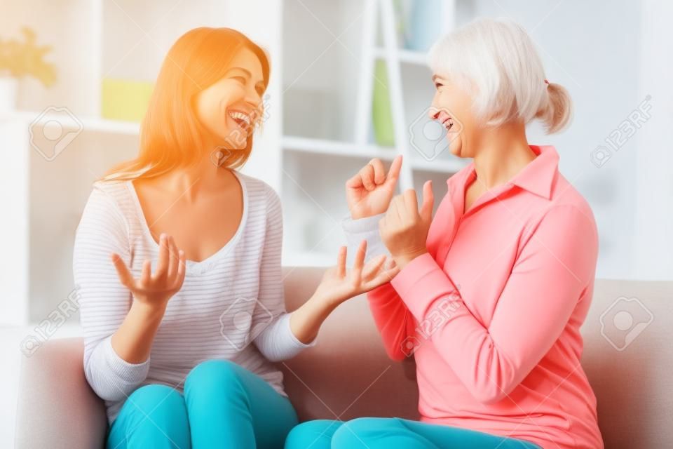 Funny dialogue. Smiling young daughter with joyful senior mother resting in living room and discussing good news while looking at each other and actively gesticulating