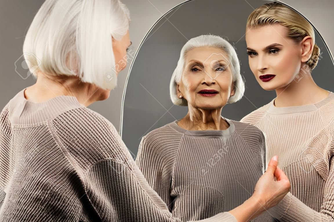 Great mood. Delighted aged woman looking into the mirror while smiling to herself