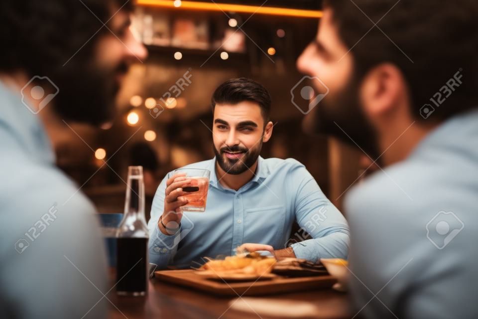 짧은 검은 머리와 수염을 가진 매력적인 멋지고 매력적인 차분한 잘생긴 남자를 쳐다보며 손에 음료수를 들고 바 테이블에서 유혹하는 친구들을 쳐다보고 있습니다