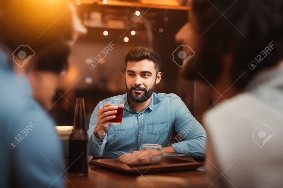 Staring at friends. Attractive nice-looking appealing calm handsome man with short dark hair and beard holding a drink in a hand and staring at his flirting friends at the bar table