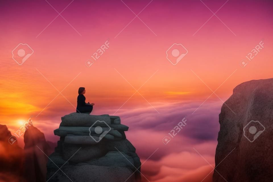 Una bella ragazza medita in una posa di loto seduto su una roccia sopra le nuvole sullo sfondo del tramonto