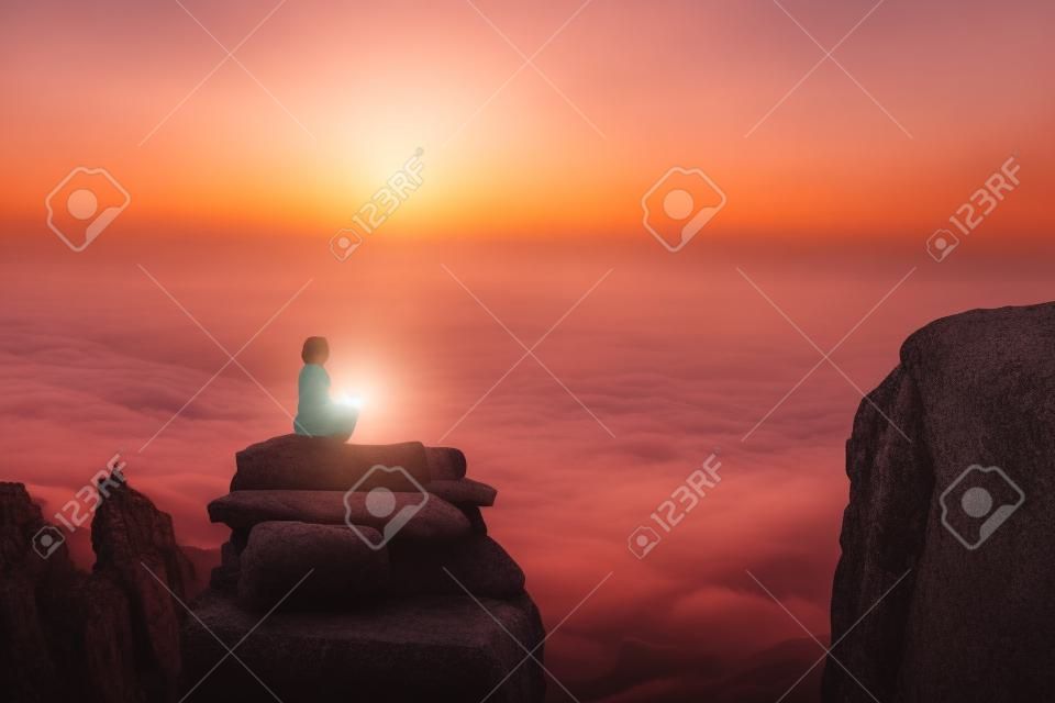 A beautiful girl meditates in a lotus pose sitting on a rock above the clouds against the backdrop of the sunset