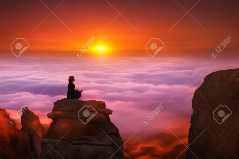 Uma bela menina medita em uma pose de lótus sentada em uma rocha acima das nuvens contra o pano de fundo do pôr do sol