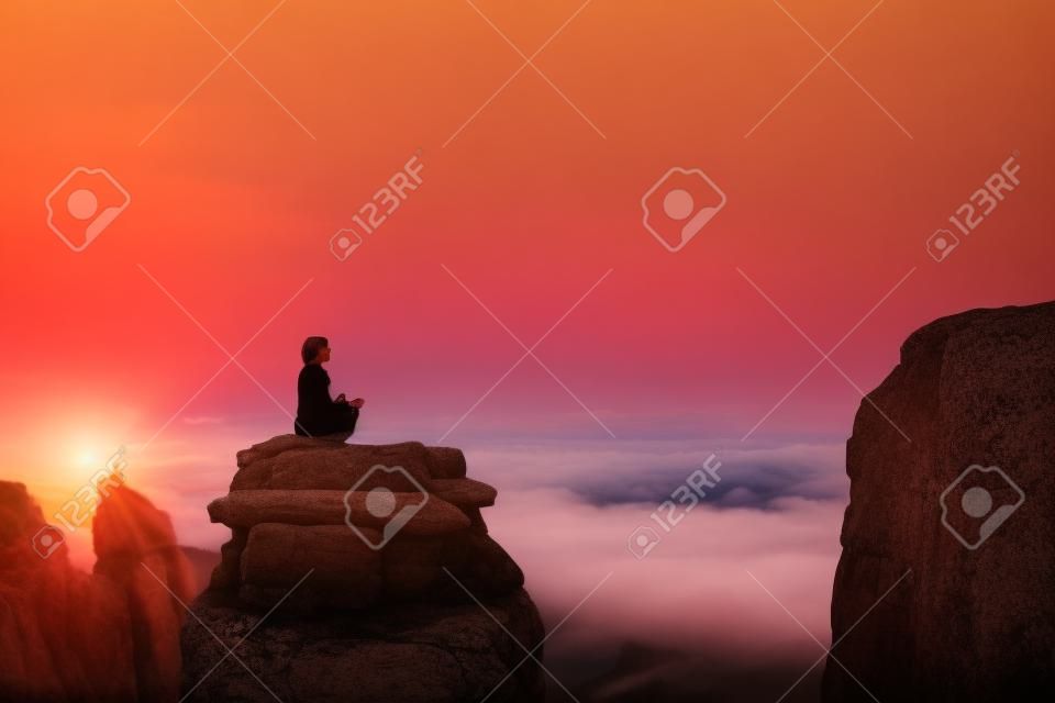 Uma bela menina medita em uma pose de lótus sentada em uma rocha acima das nuvens contra o pano de fundo do pôr do sol