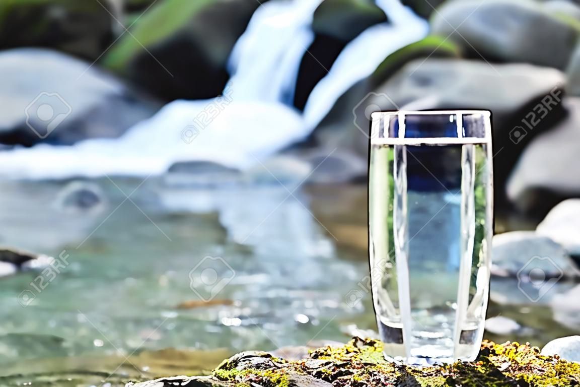 Um vidro de vidro transparente com água de montanha potável fica na pedra de musgo no raio de sol em um fundo de um rio de montanha de gelo limpo. O conceito de beber água mineral de montanha bebendo e a produção de água mineral potável ambientalmente amigável