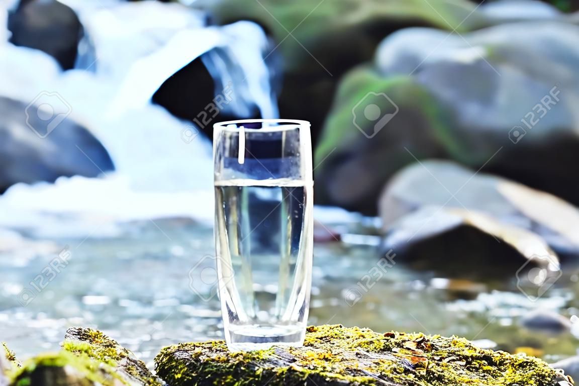 Um vidro de vidro transparente com água de montanha potável fica na pedra de musgo no raio de sol em um fundo de um rio de montanha de gelo limpo. O conceito de beber água mineral de montanha bebendo e a produção de água mineral potável ambientalmente amigável