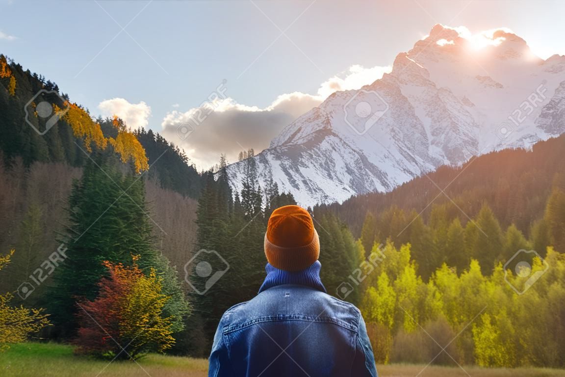 Back view of a man in a knitted hat and denim jacket against the backdrop of a green dense forest and snow-capped high mountains