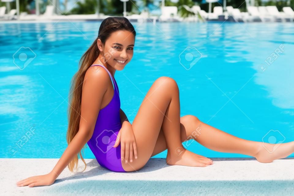 Schönes junges Mädchen im lila Badeanzug, der am Pool sitzt und zur Kamera lächelt