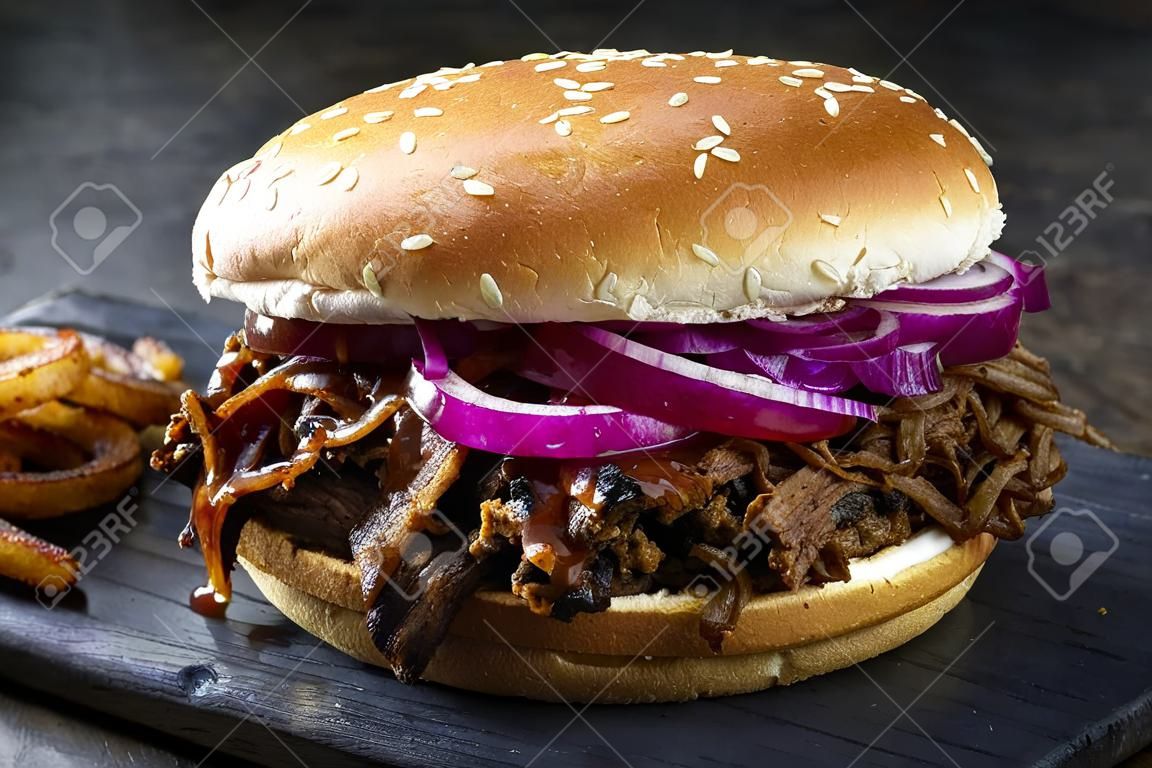 Traditioneller Barbecue Pulled Beef Burger mit Carolina BBQ Sauce, Gurke und Zwiebelringen als Nahaufnahme auf einem verbrannten Holzbrett