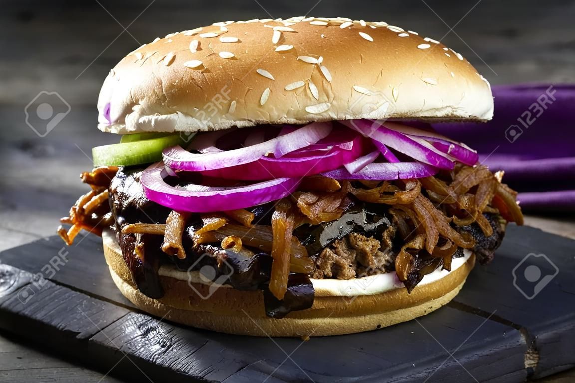 Traditional barbecue pulled beef burger offered with Carolina BBQ sauce, gherkin and onion rings as closeup on a burnt wooden board