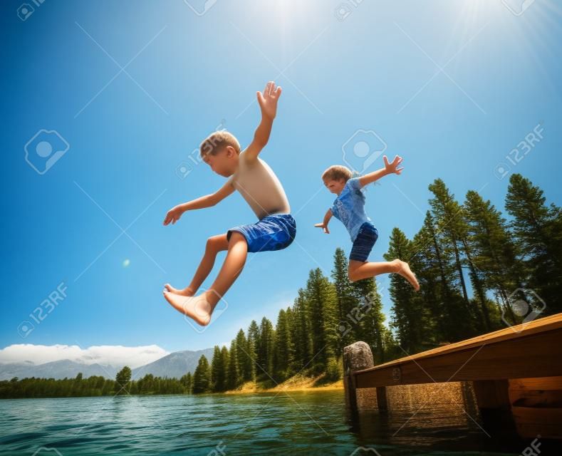 Niños saltando del muelle a un hermoso lago de montaña. Divirtiéndose en unas vacaciones de verano en el lago con amigos