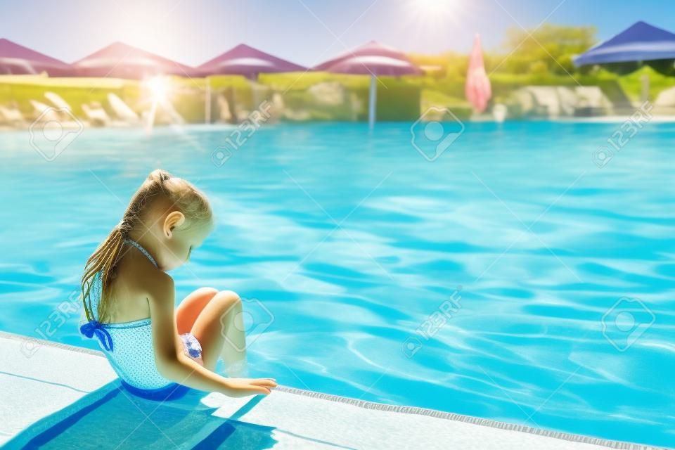 A lovely little girl sits on the side of the pool with her feet in the water enjoying the sun